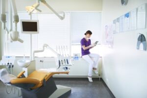 A dentist in scrubs sitting in an exam room looking at a tablet.